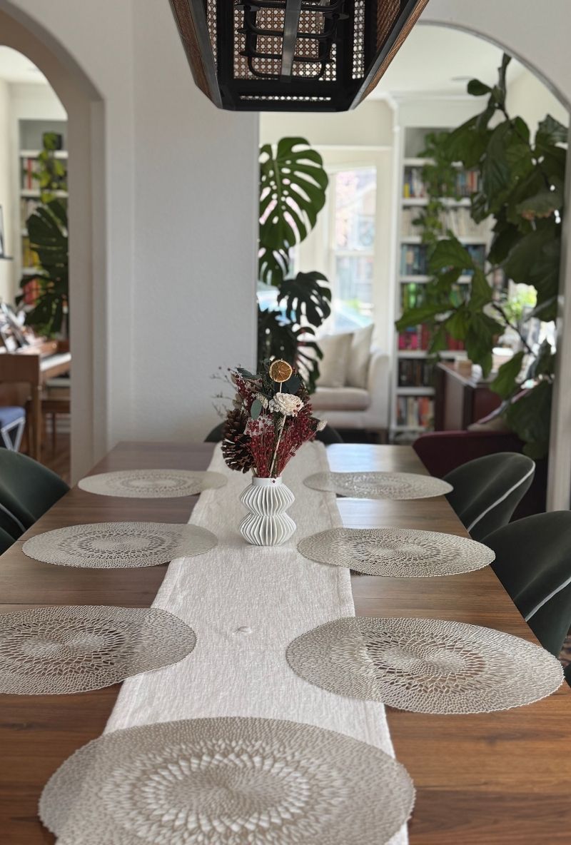 A wooden dining table set with a white runner, intricate silver placemats, and a vase with a dried floral arrangement.